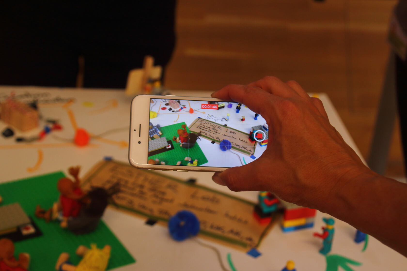 Eine Hand hält ein Smartphone mit aktivem Bildschirm im Bildaufnahmemodus. Auf dem Bildschirm des Smartphones ist das Objekt der Aufnahme zu sehen: eine Lego-Landschaft und ein Zettel, auf dem Esther Lab steht.