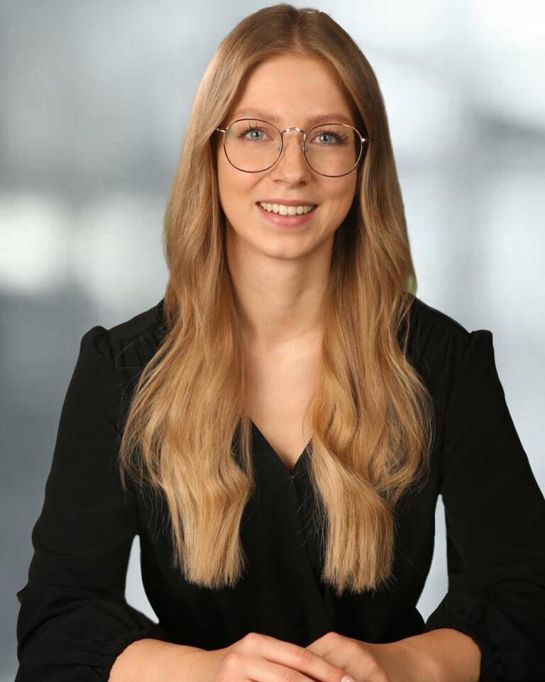 Portraitbild von Julia Pusterhofer mit langen blonden gewellten Haaren, Brille, schwarzer Bluse mit grauem Hintergrund