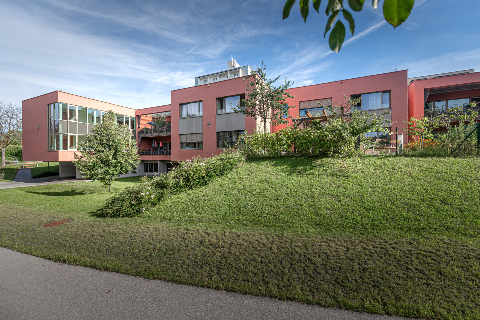 Außenansicht des roten Gebäudes im Pflegewohnheim Aigner Rollett am Rosenhain mit großer Grünfläche und Bäumen