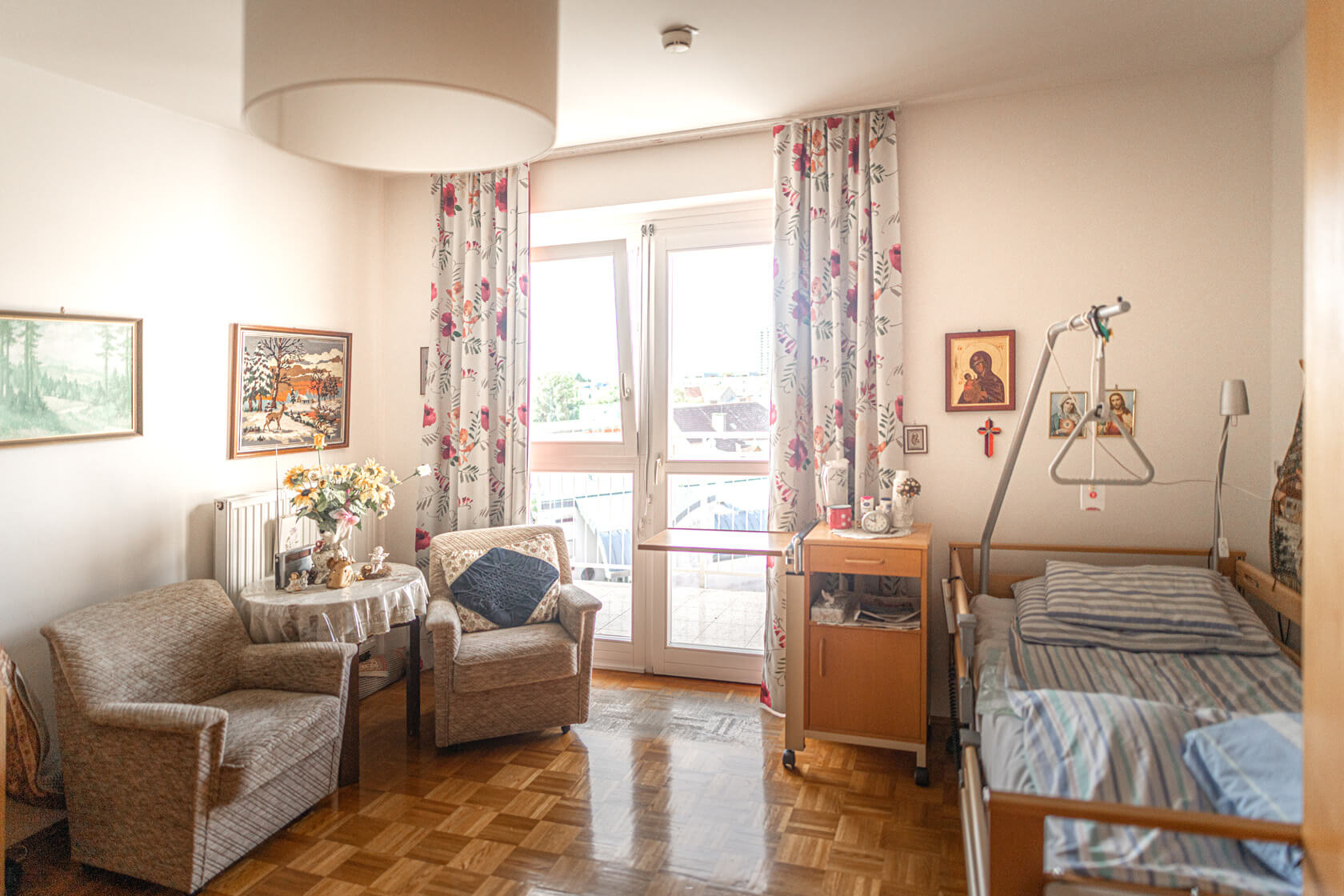 Bewohner:innenzimmer im Pflegewohnheim Aigner Rollett am Rosenhain mit Sitzecke, Bett, Parkettboden und verschiedenen Bildern an der Wand
