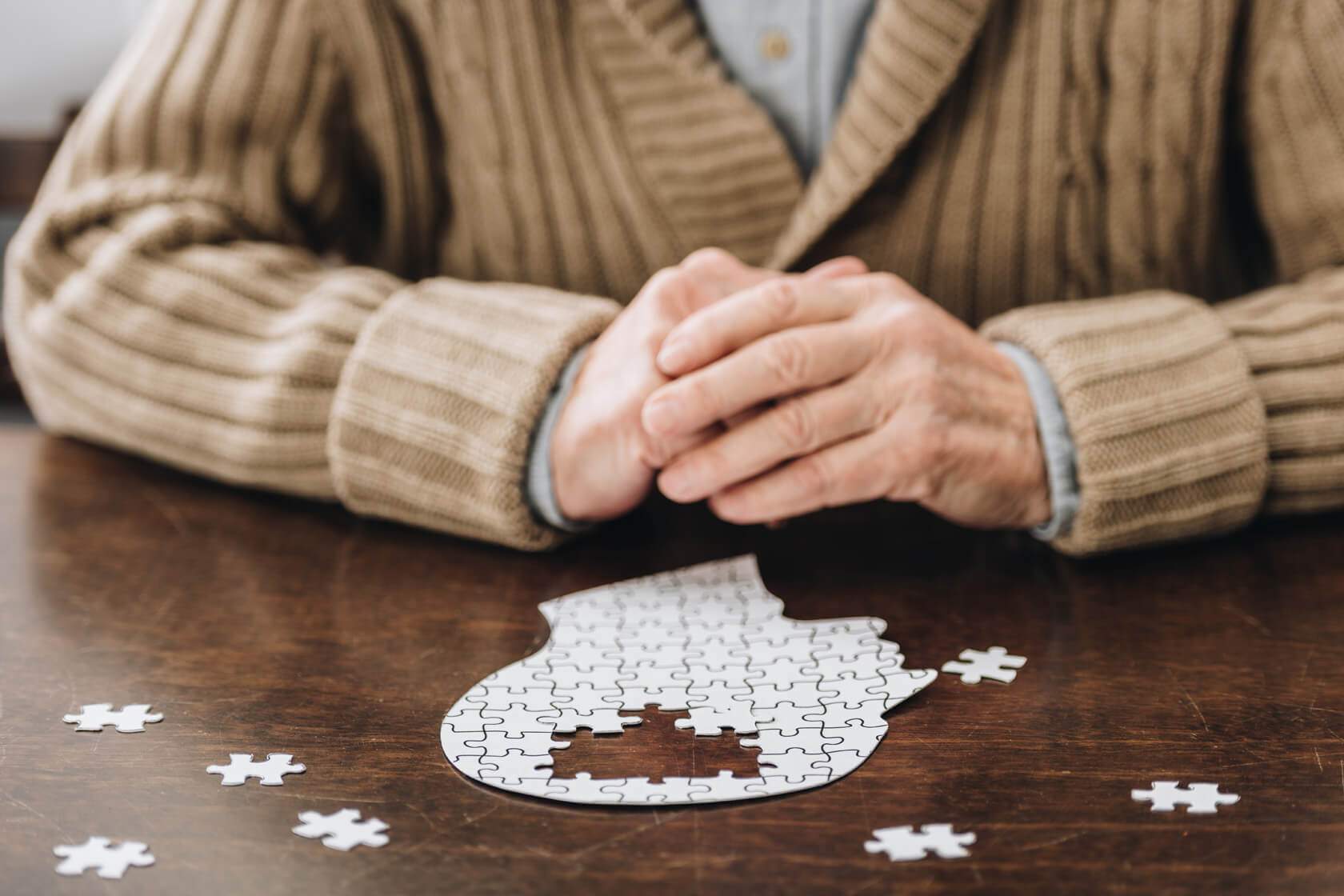 Ein Puzzle in Form eines Kopfes liegt halb fertig auf einem Tisch und davor sitzt eine ältere Person, die ihre Hände verschränkt hat.