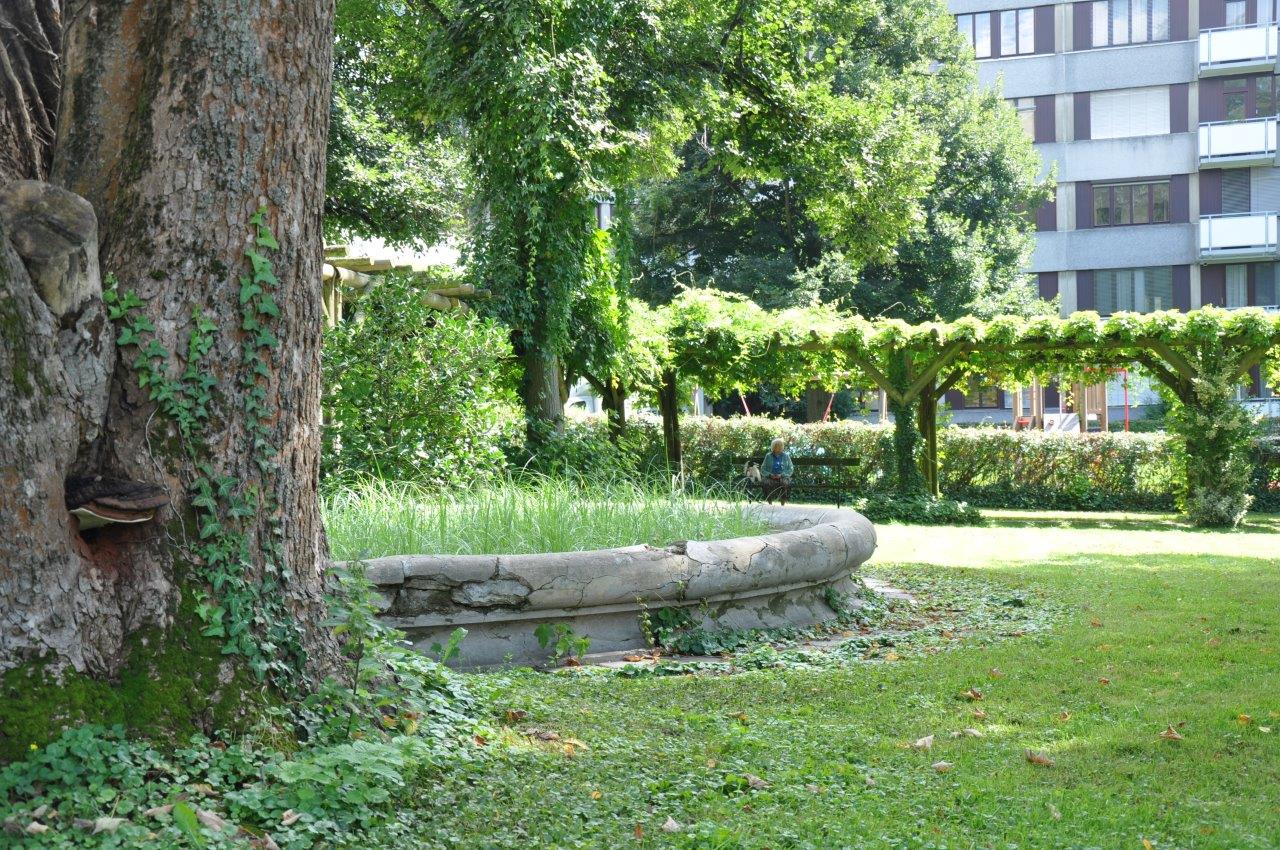 Ein großer Baum steht neben einem Steinbrunnen und im Hintergrund ist eine Frau zu sehen die sich entspannt auf eine schattige Bank gesetzt hat.