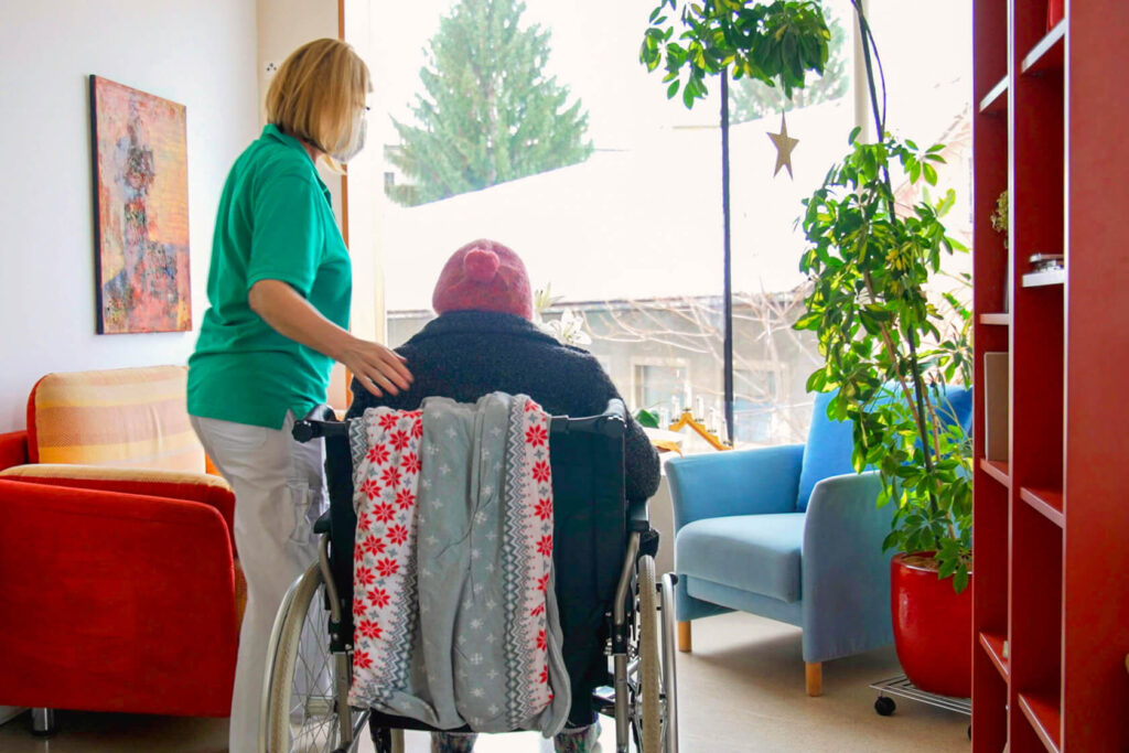 Stationsleiterin mit Patientin im Rollstuhl in Bücherecke im Albert Schweitzer Hospiz. Beider Personen sind von hinten zu sehen.