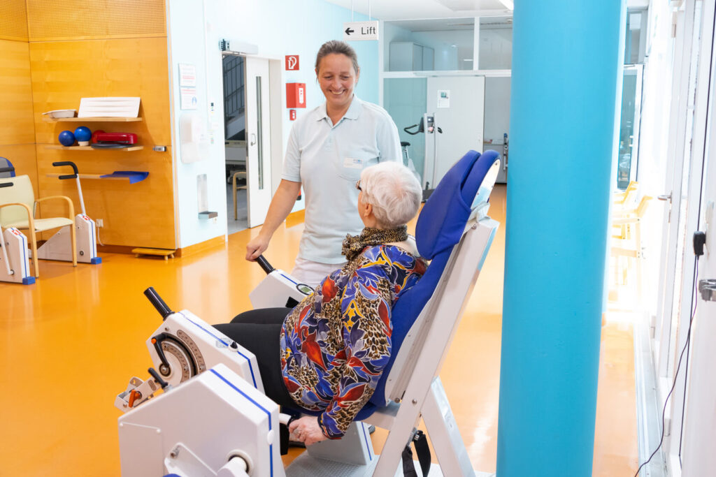 Eine Physiotherapeutin steht vor einer geriatrischen Patientin, die auf einem Trainingsgerät sitzt.