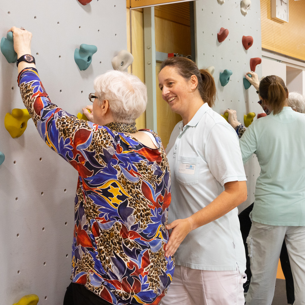 Patientin in der Tagesklinik im Trainingsbereich beim Training auf Kletterwand. Daneben steht eine Therapeutin und unterstützt sie bei den Übungen.