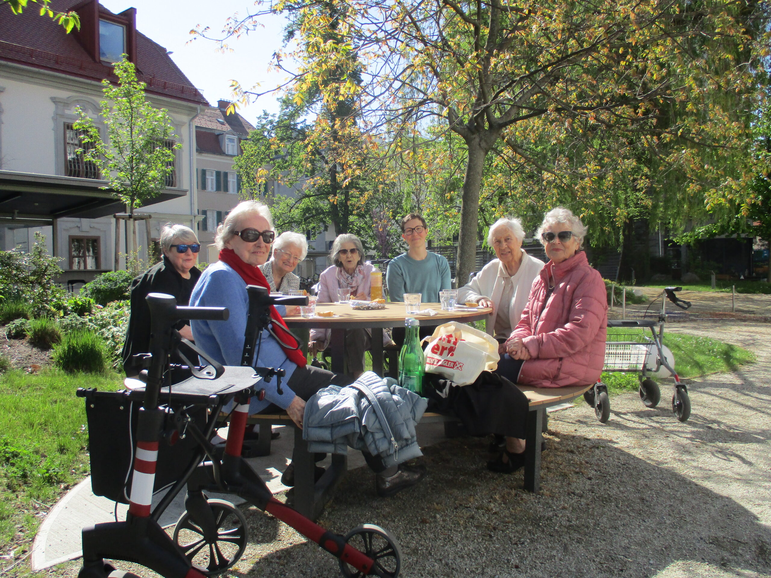 Sechs Senior:innen sitzen mit einer Betreuerin an einem runden Tisch und picknicken.