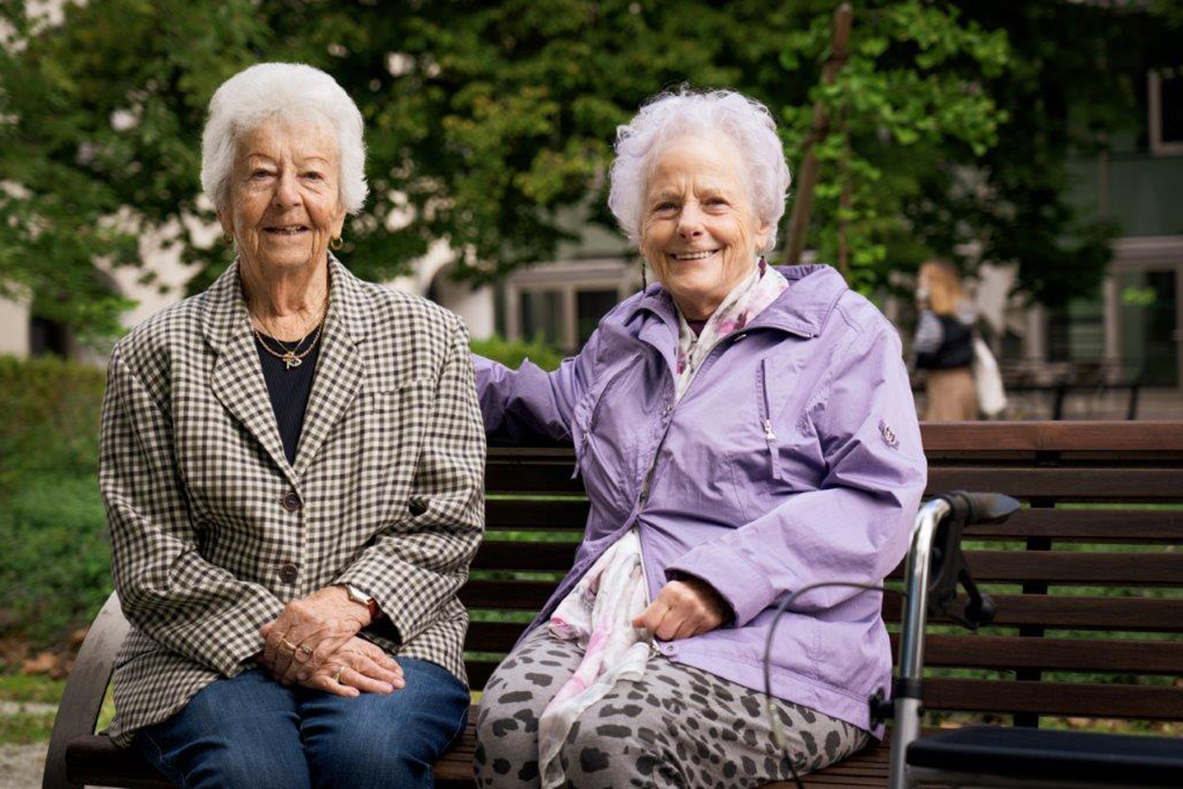 Zwei Seniorinnen mit Rollator sitzen im Freien auf einer Bank.