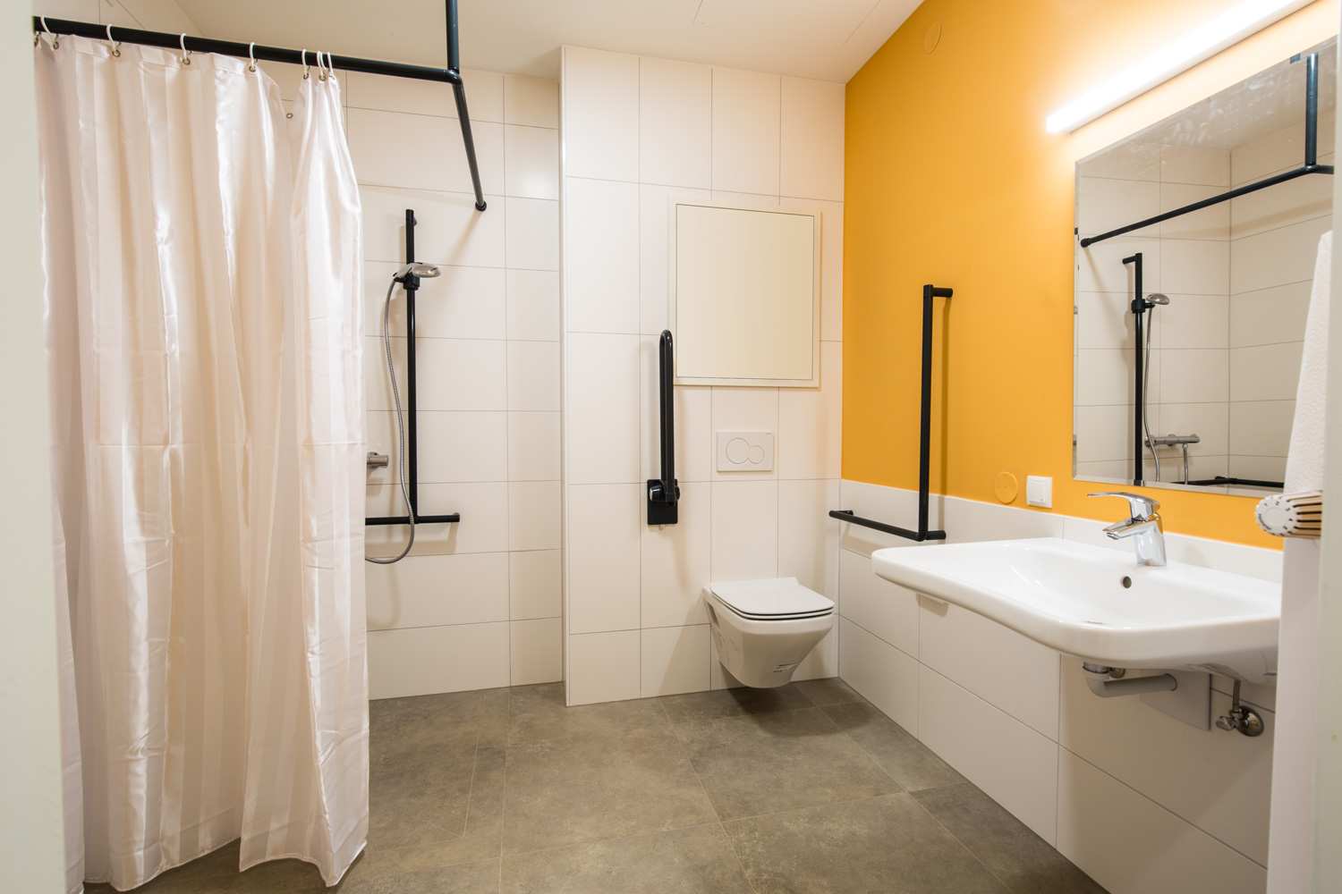 Ein großes Badezimmer mit einer Dusche mit Duschvorhand, einem Klo mit Stangen an der Wand und einem großen Wachbecken mit Spiegel darüber.