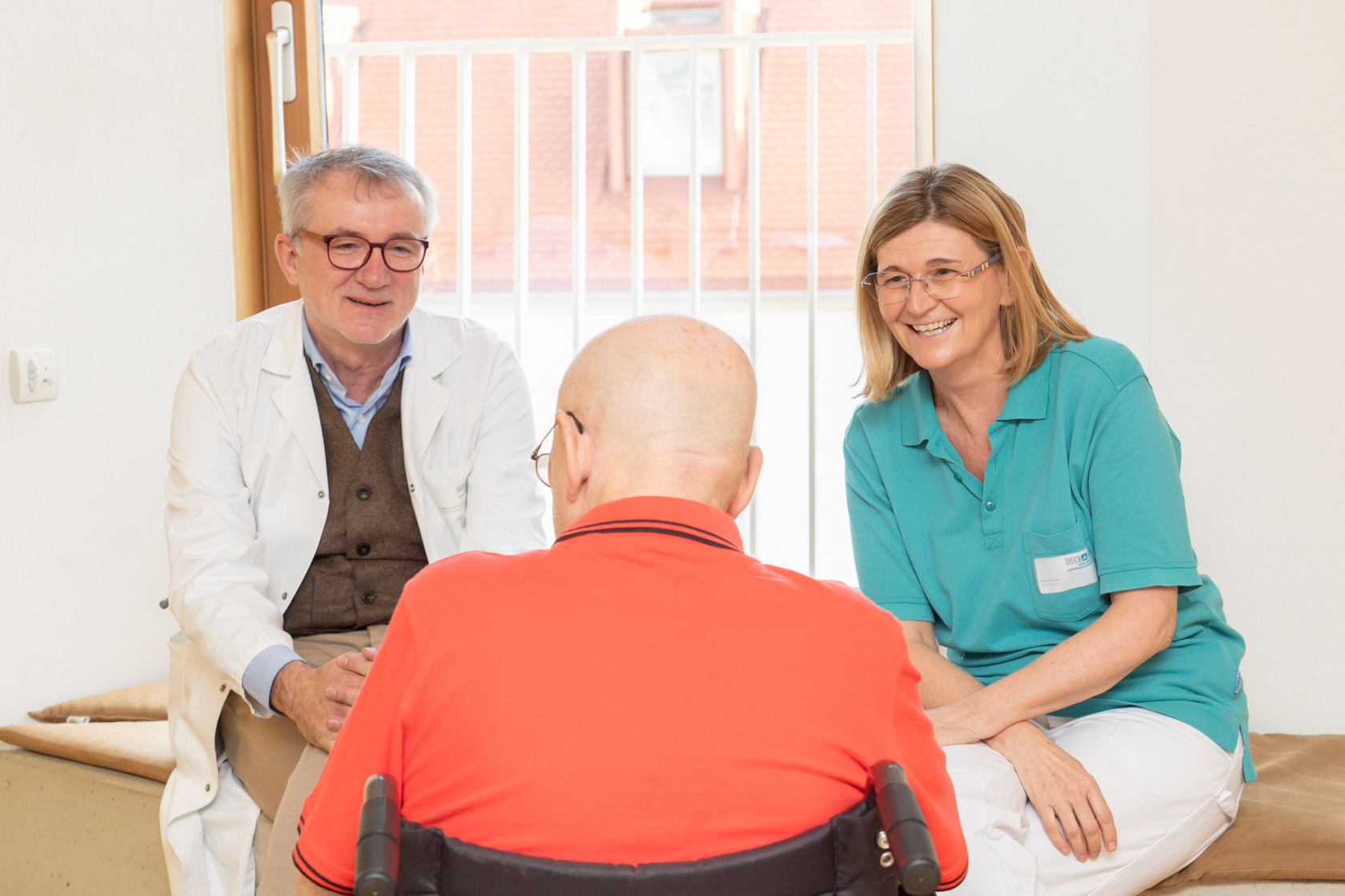 Arzt und Stationsleitung im Albert Schweitzer Hospiz sitzen am Fenster eines Patientenzimmers und sprechen mit einem Hospizpatienten, der im Rollstuhl sitzt