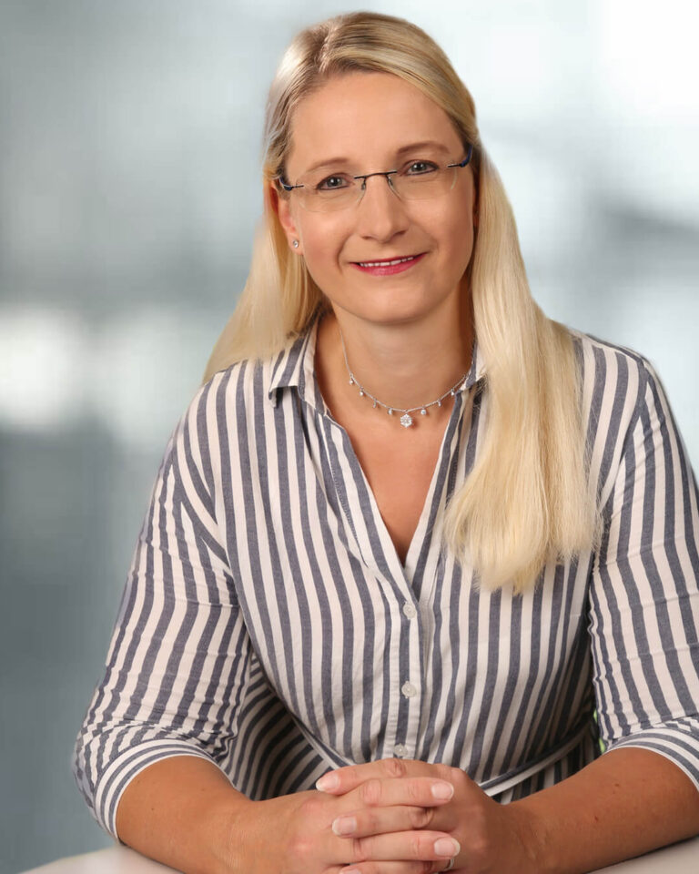 Portraitfoto von Eva Ramsenthaler mit langen blonden Haaren, rahmenloser Brille, Kette und gestreifter Bluse.