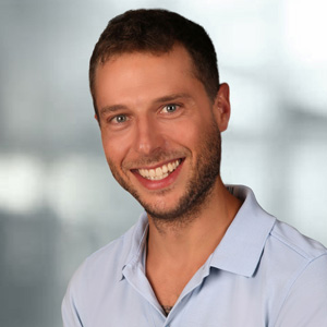 Portraitbild von Ranegger Matthias mit braunen kurzen Haaren, grauem Hintergrund, blauem T-Shirt, Bart und schönem Lächeln