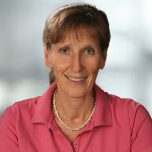 Portraitbild von Reitbauer Marianne mit braunen zusammengebundenen Haaren, grauem Hintergrund, pinkem T-Shirt, Halskette und schönem Lächeln