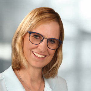 Portraitbild von Claudia Seidl mit blonden Haaren, grauem Hintergrund, blauem T-Shirt, Halskette und schönem Lächeln