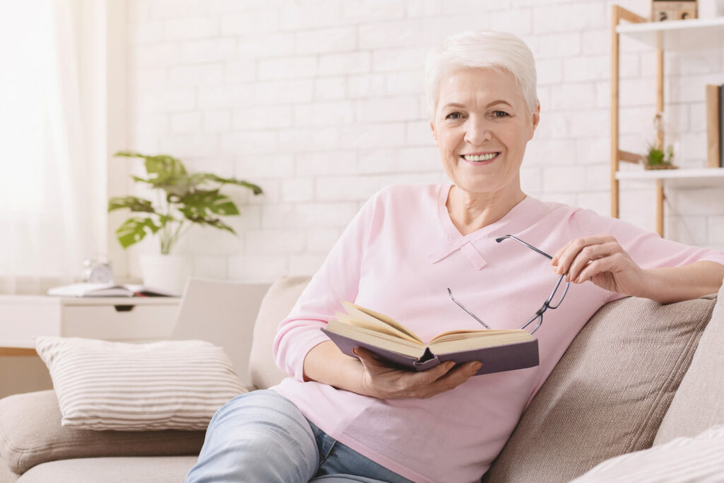 Eine Seniorin sitzt auf einer Couch in angenehmer Atmosphäre, hält eine Brille und ein Buch in der Hand und lächtelt in die Kamera.