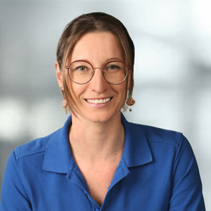 Portraitbild von Stöckl Anita mit braunen zusammengebundenen Haaren, grauem Hintergrund, blauem T-Shirt und schönem Lächeln