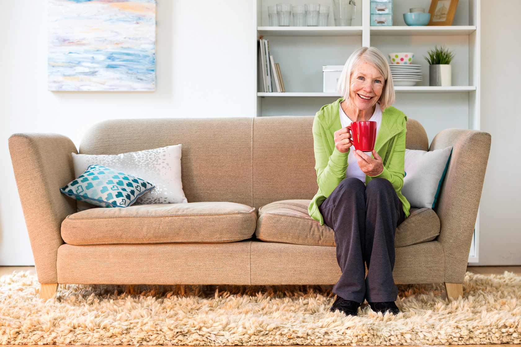 Seniorin sitzt mit roter Kaffeetasse auf Couch im Betreuten Wohnen und lächelt
