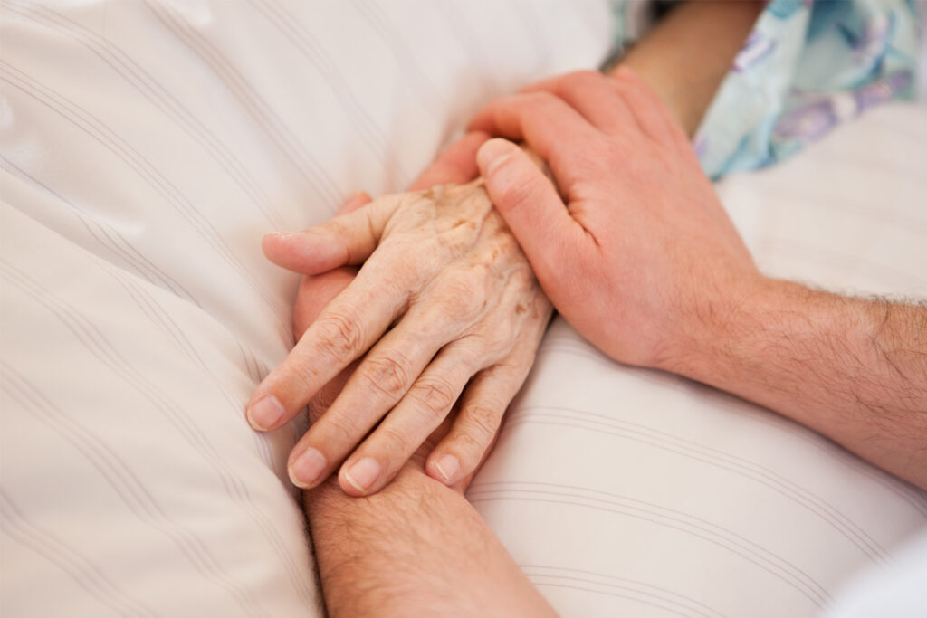 Zwei Hände halten die Hand einer älteren Person die im Bett liegt.