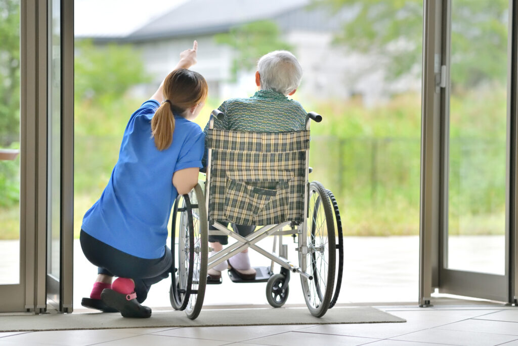 Eine Pflegerin hockt neben einem älteren Mann im Rollstuhl und zeigt mit ihrem Finger nach draußen in die Ferne.