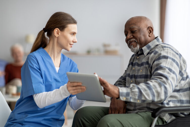 Eine Pflegerin hält ein Tablet in der Hand und hilft einem älteren Herrn im Rollstuhl dabei es zu verwenden.