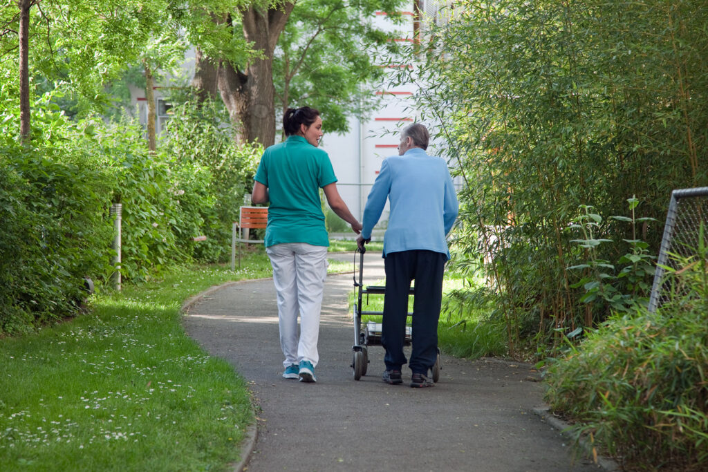 Pflegerin mit türkisem Shirt geht im Demenzgarten mit Senior mit Rollator spazieren