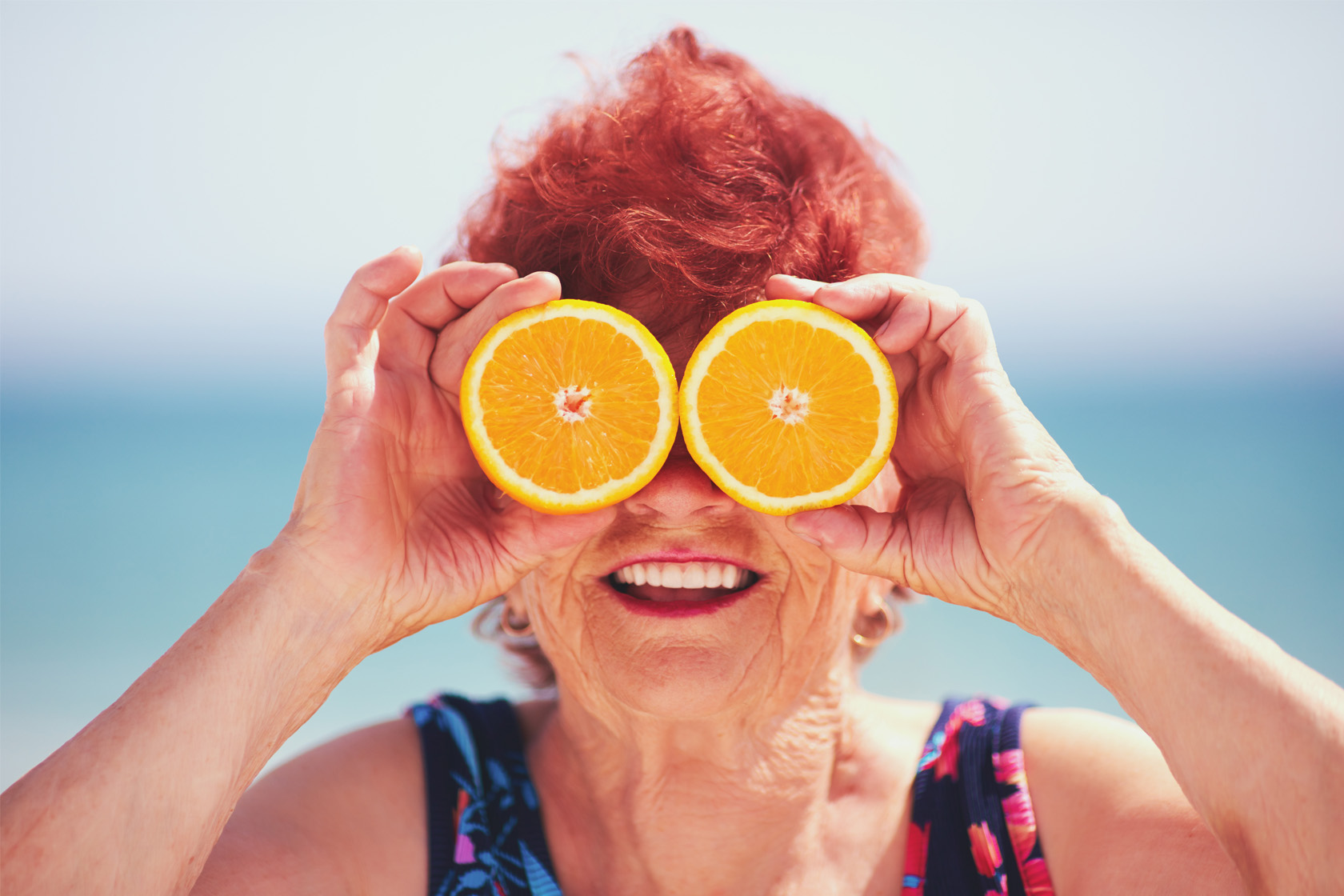 Eine ältere Dame steht am Meer und hält sich lächelnd zwei Orangenscheiben vor die Augen.