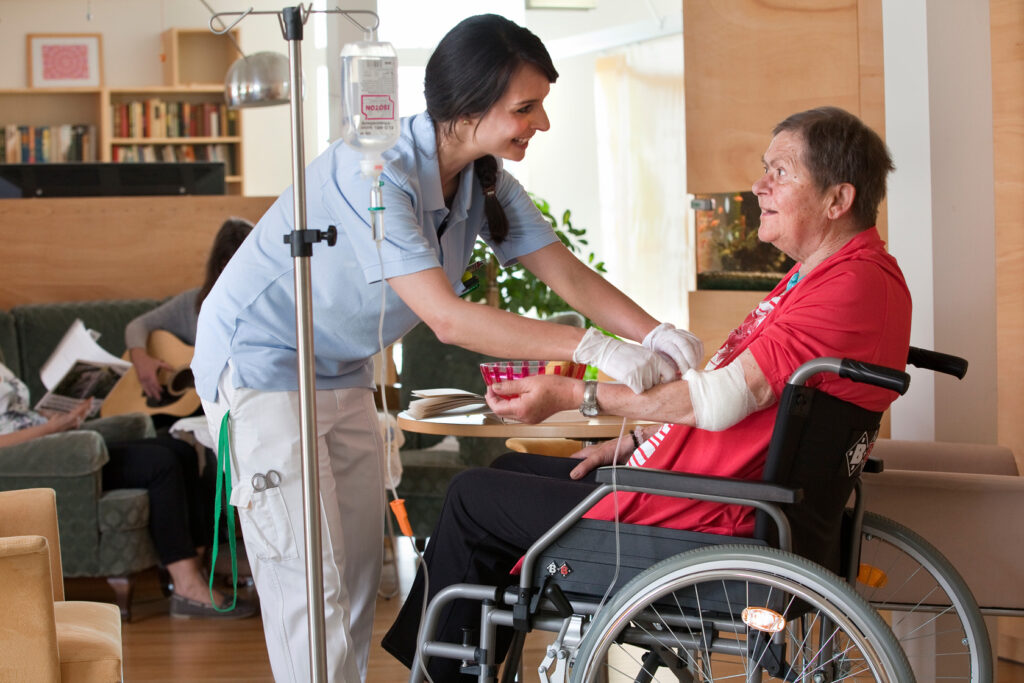 Pflegefachkraft mit blauen T-Shirt und weißer Hose legt einer Bewohnerin im Rollstuhl einen Zugang