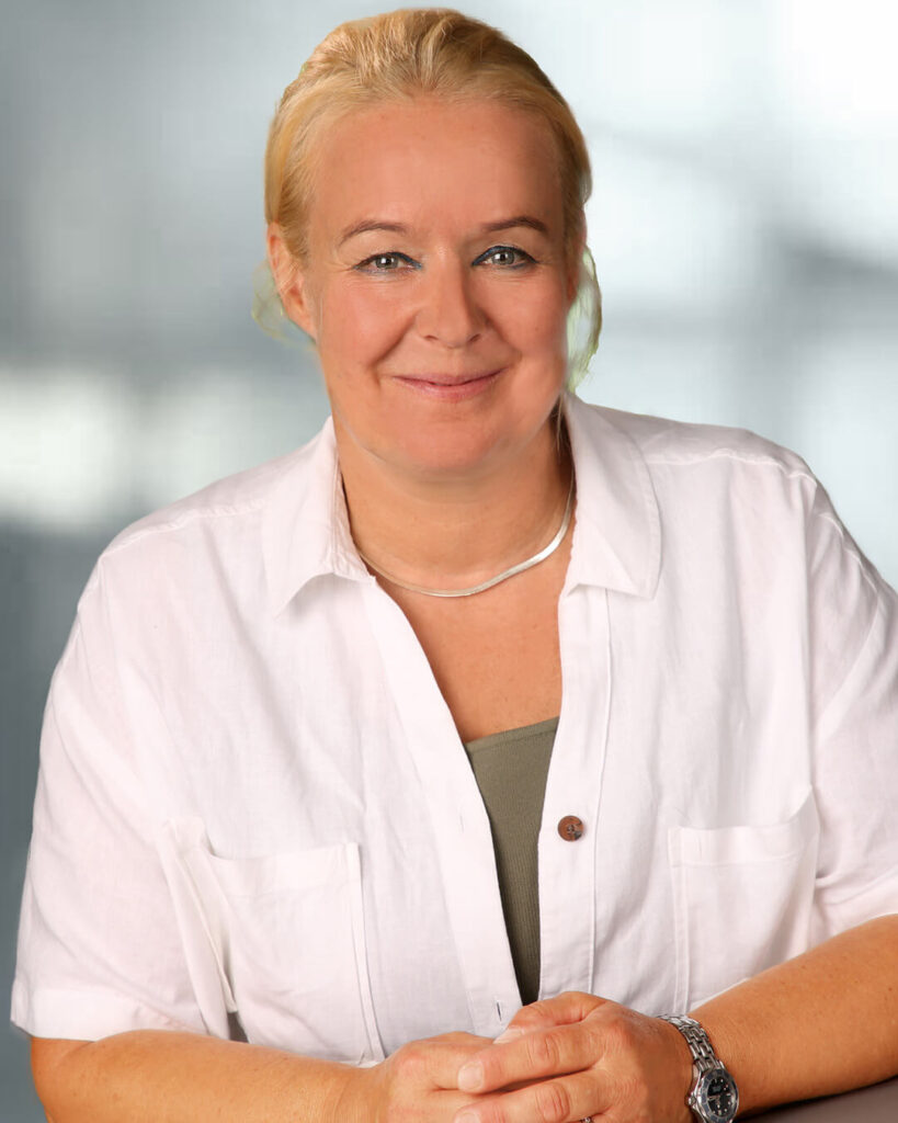 Portraitfoto von Karin Marx-Lederhaas mit blonden zusammengebundenen Haaren, weißer Bluse und grünem Shirt.