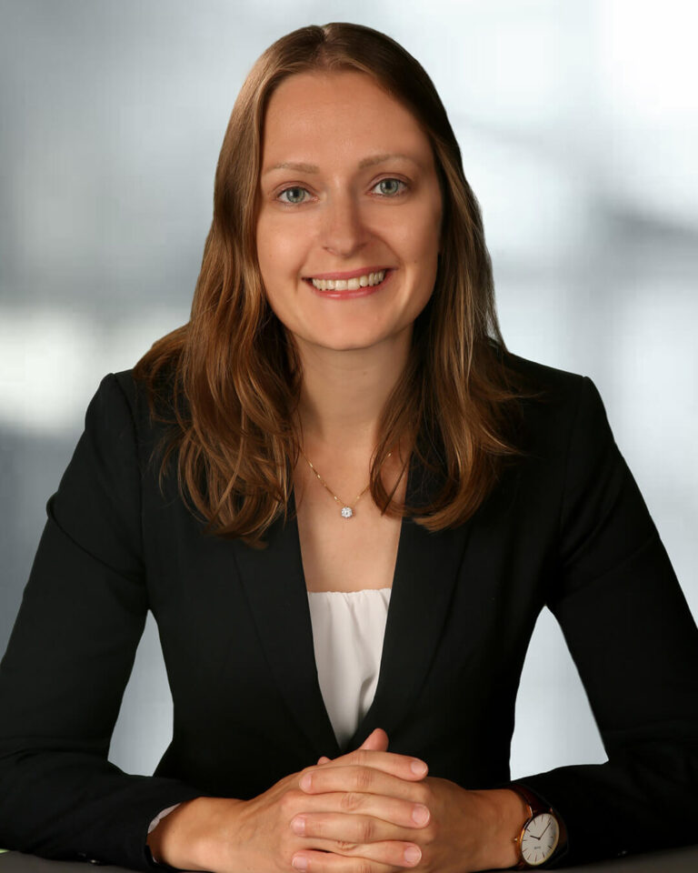 Portraitbild Verena Matz mit braunen Haaren, weißem Shirt und schwarzem Blazer und grauem Hintergrund