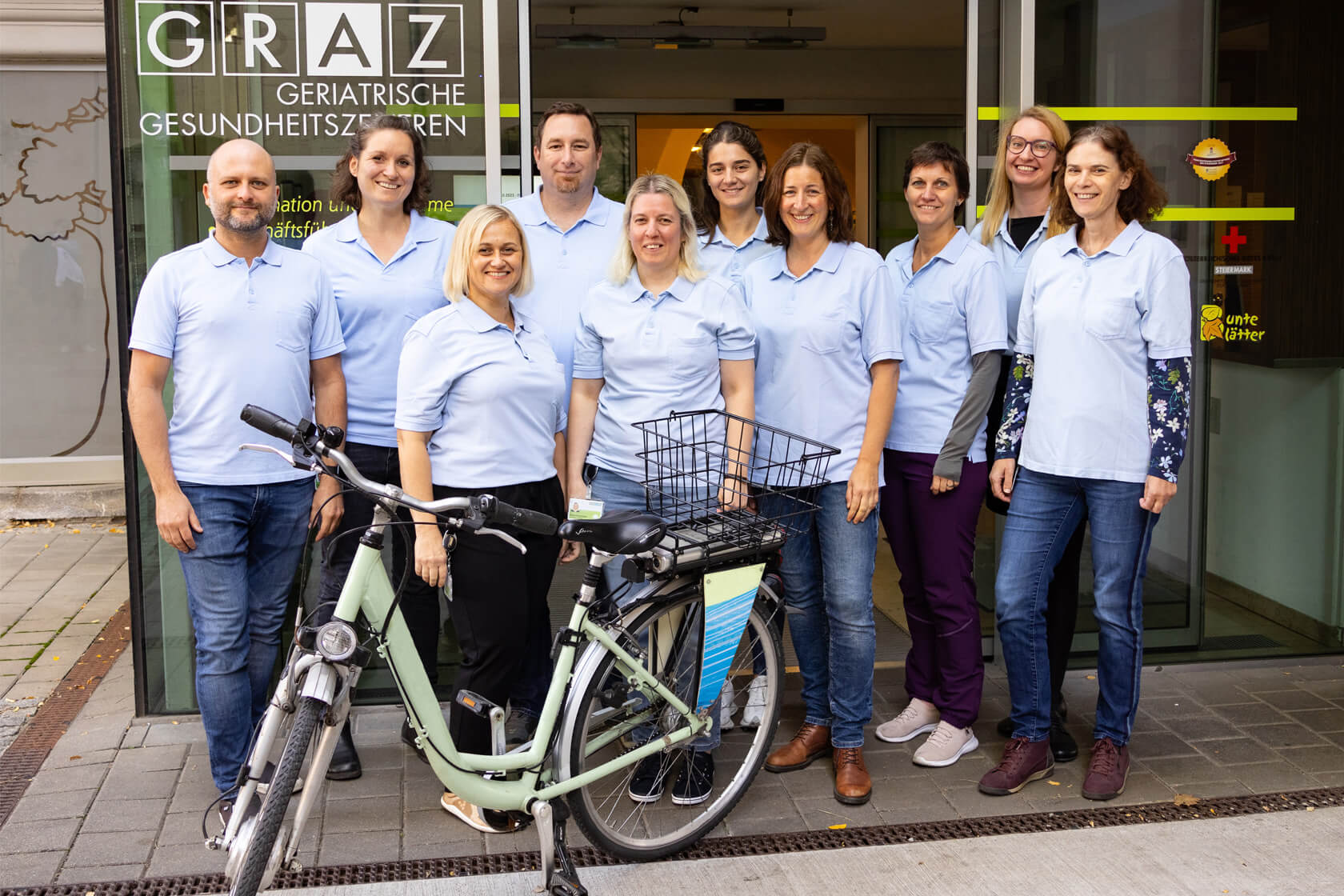 Gruppenfoto des Teams der mobilen Remobilisation vor dem Eingangsbereich der Geriatrischen Gesundheitszentren mit blauen T-Shirts und e-Bike im Vordergrund