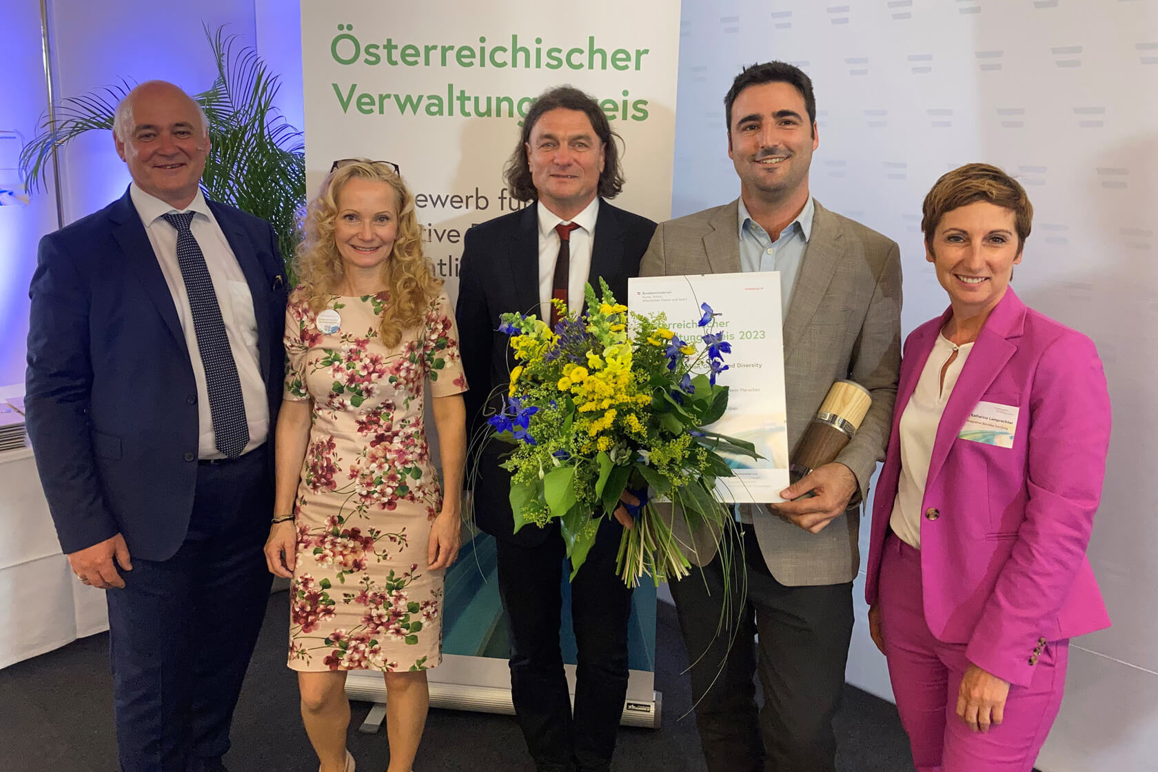 Fünf Menschen stehen lächelnd nebeneinander. Der Mann in der Mitte hält einen Blumenstrauß und ein Zertifikat in der Hand mit dem der Verwaltungspreis 2023 verliehen wurde.