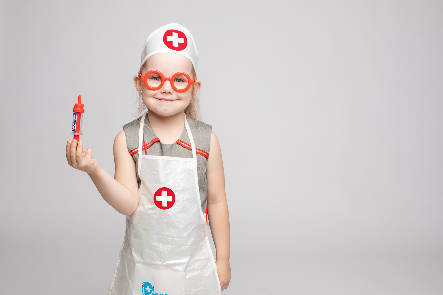 Ein kleines Mädchen steht in einem Pfleger-Kostüm vor einer weißen and und hält lächelnd eine Spielzeugspritze in der Hand.