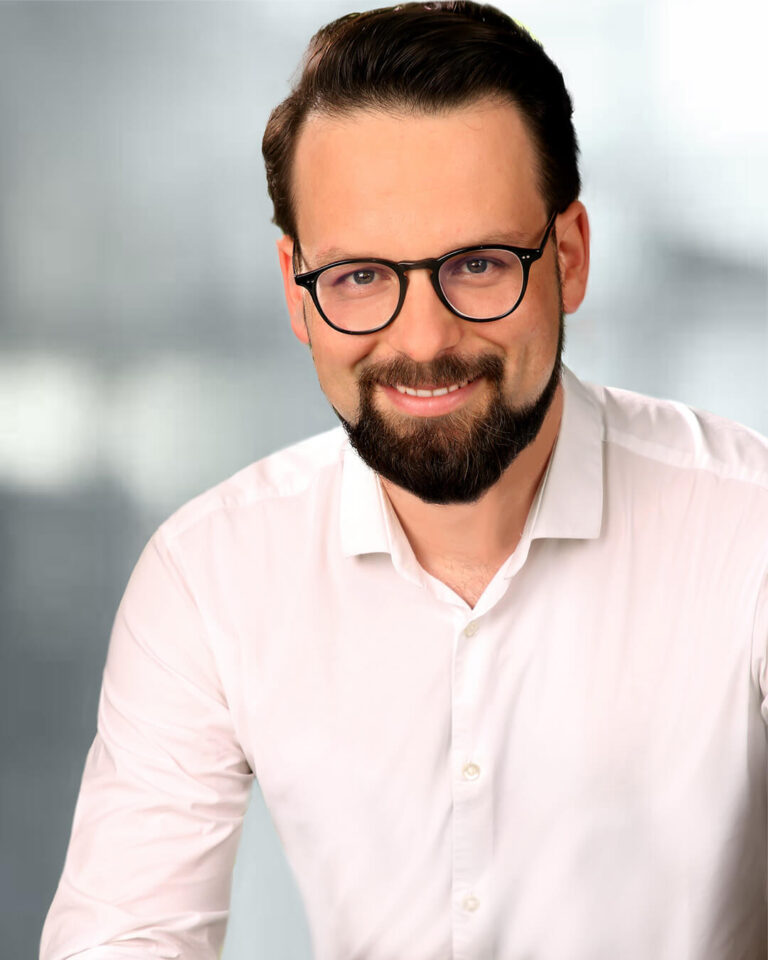 Portraitfoto von Christoph Kocher mit Brille, dunklen Haaren, weißem Hemd und grauem Hintergrund