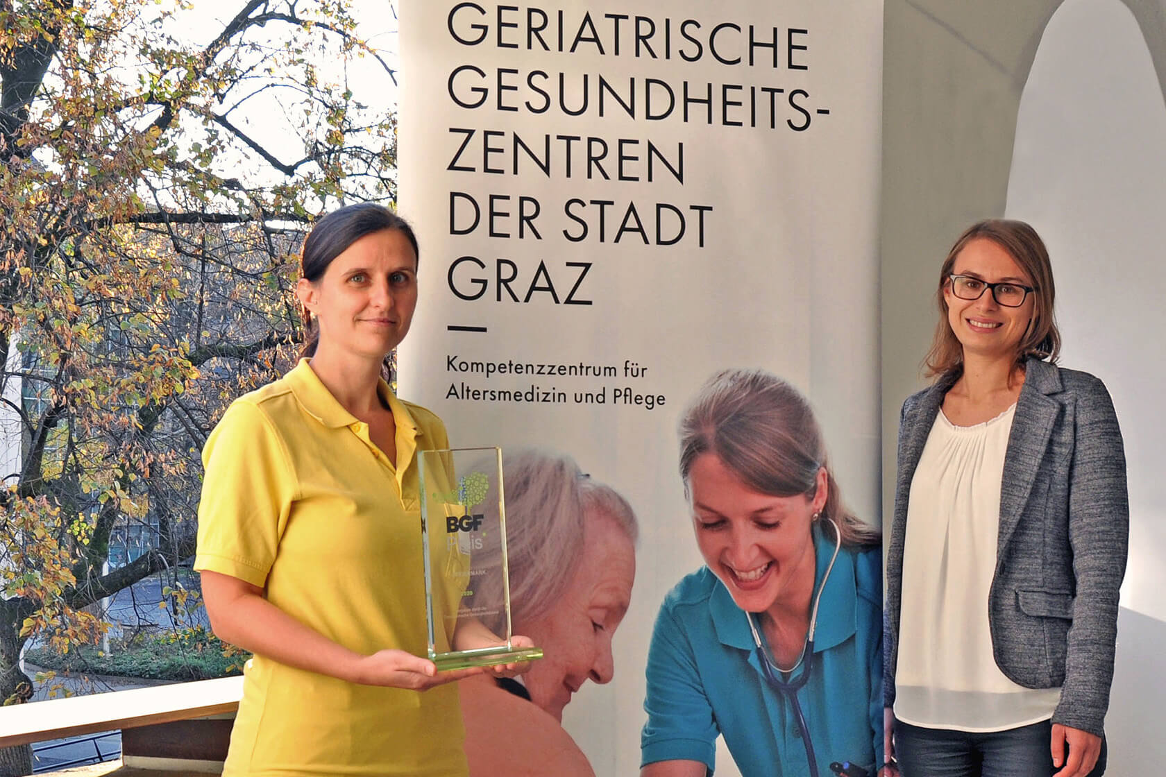 Zwei GGZ Mitarbeiterinnen vor den GGZ Roll up mit dem Preis für Betriebliche Gesundheitsförderung in den Händen
