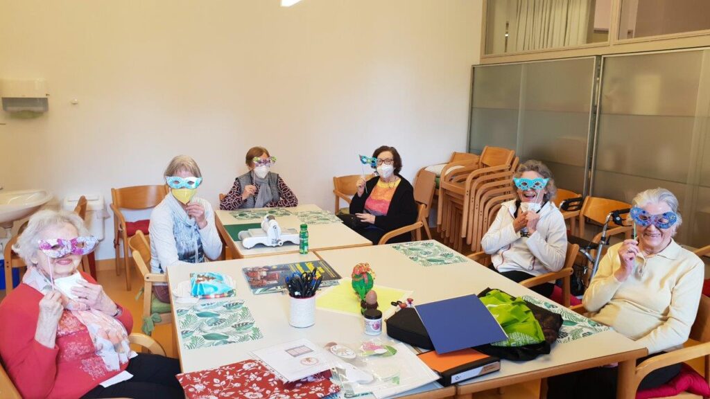 Tagesgäste sitzen an einem Tisch zu Fasching im Kreis und halten Masken vor ihr Gesicht