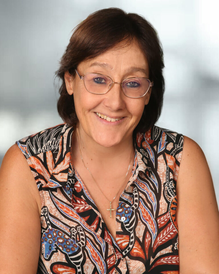 Portraitbild von Renate Bauer mit dunklem schulterlangem Haar, grauem Hintergrund bunter, ärmelloser Bluse und Brille