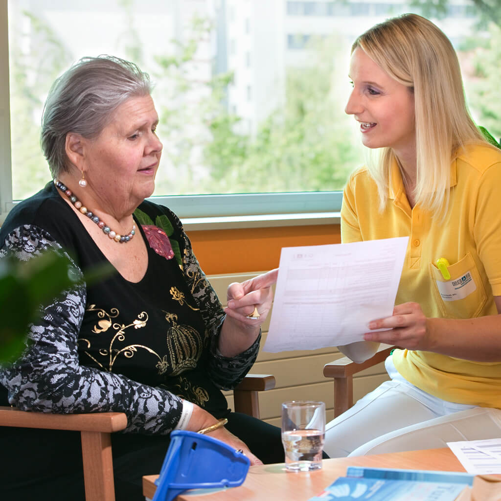 Pflegerin mit gelben T-Shirt und blondem Haar erklärt Patientin im Aufenthaltsbereich der Station der Albert Schweitzer Klinik ein Formular.