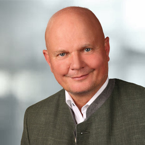 Portraitfoto von Pflegedienstleiter Jörg Hohensinner mit weißem Hemd, Sakko und grauem Hintergrund