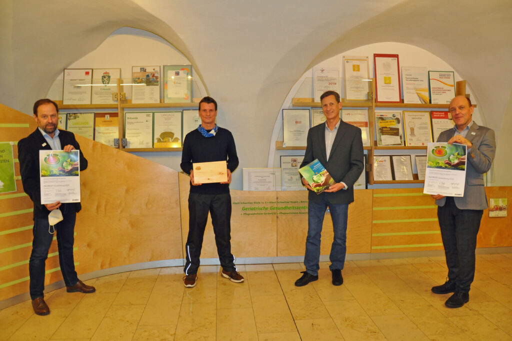GGZ Mitarbeiter und Geschäftsführer vor einer Vielzahl an gerahmten Urkunden halten Auszeichnung und Urkunde für den Ökoprofit Preis in den Händen