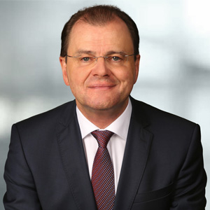 Portraitfoto von Primar Walter Schippinger mit weißem Hemd, Krawatte und schwarzem Sakko und grauem Hintergrund