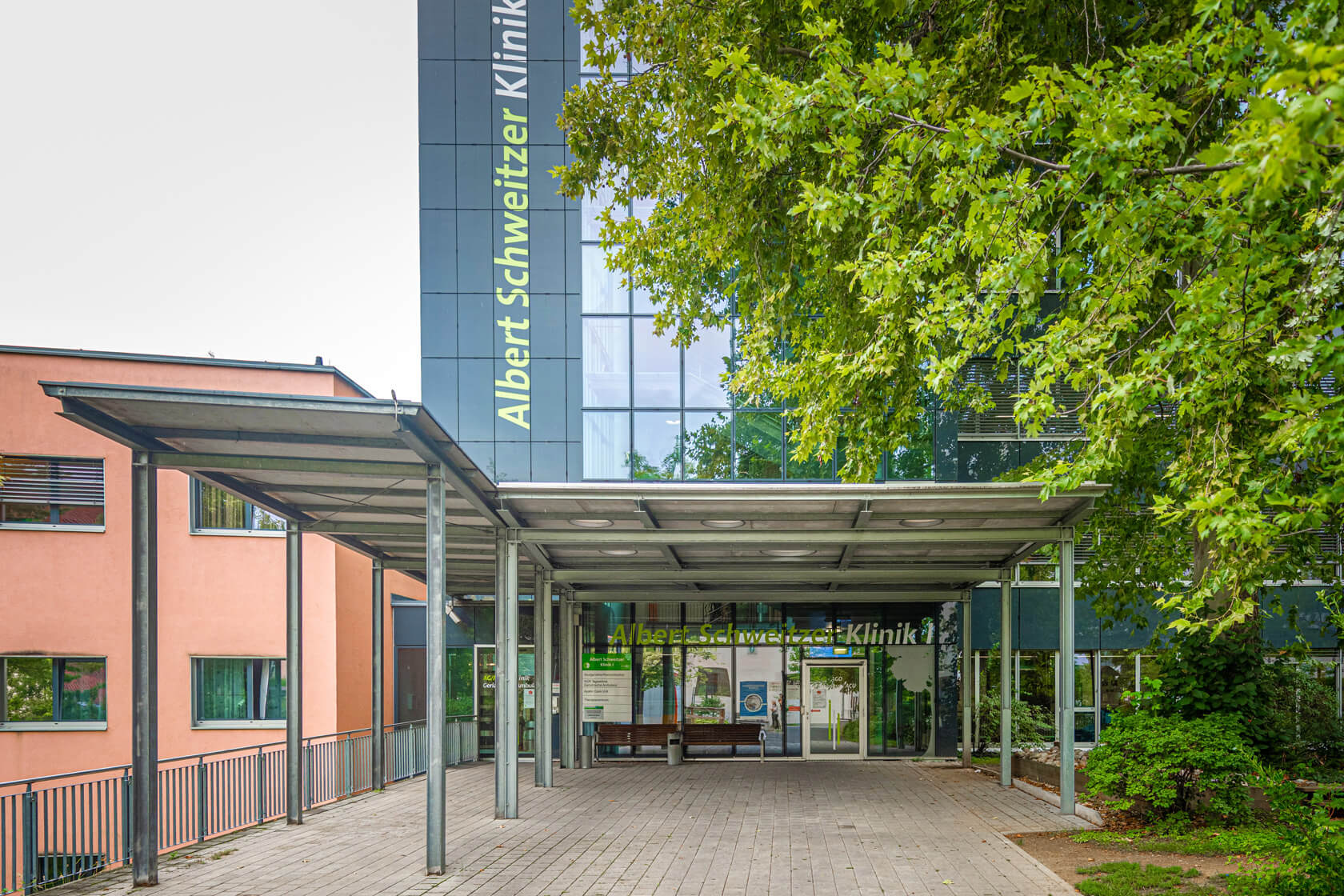Außenaufnahme des Eingangs eines Gebäudes, auf dem Albert Schweitzer Klinik 1 steht. Der Eingangsbereich ist überdacht und von Bäumen umgeben.