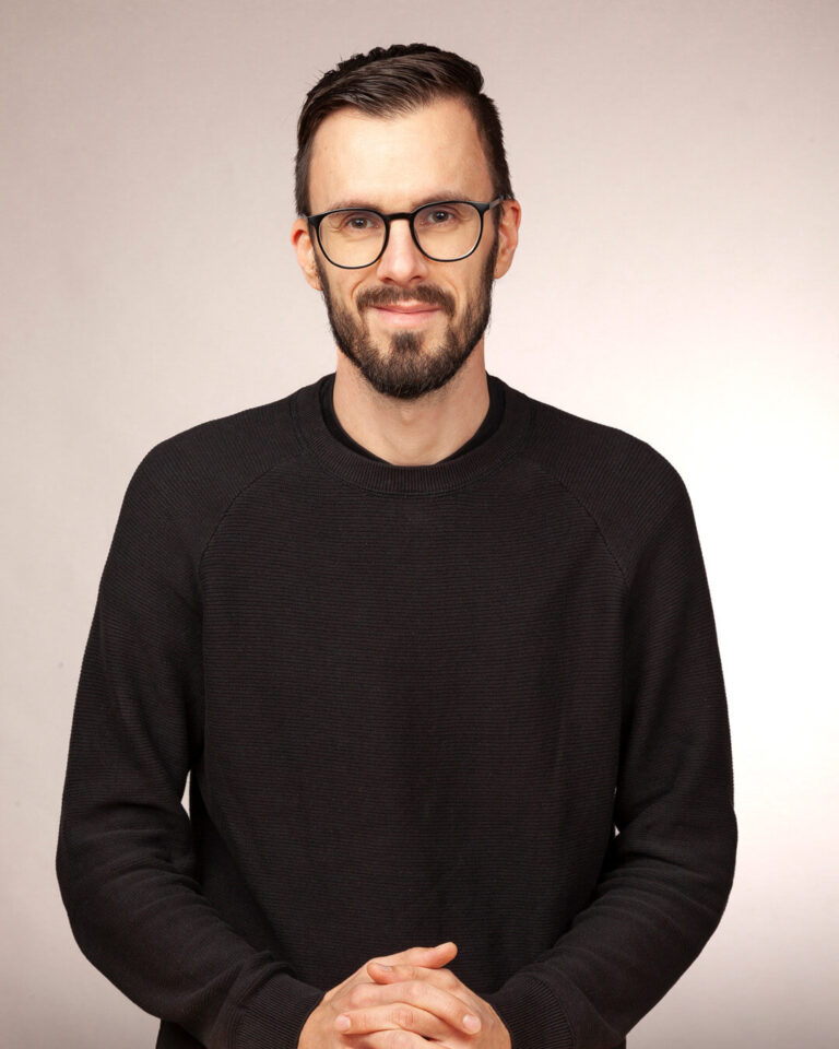 Ein Mann mit dunkelbraunen kurzen Haaren, Brille und Dreitagebart, gekleidet in einem schwarzen Pullover, blickt offen in die Kamera und lächelt.