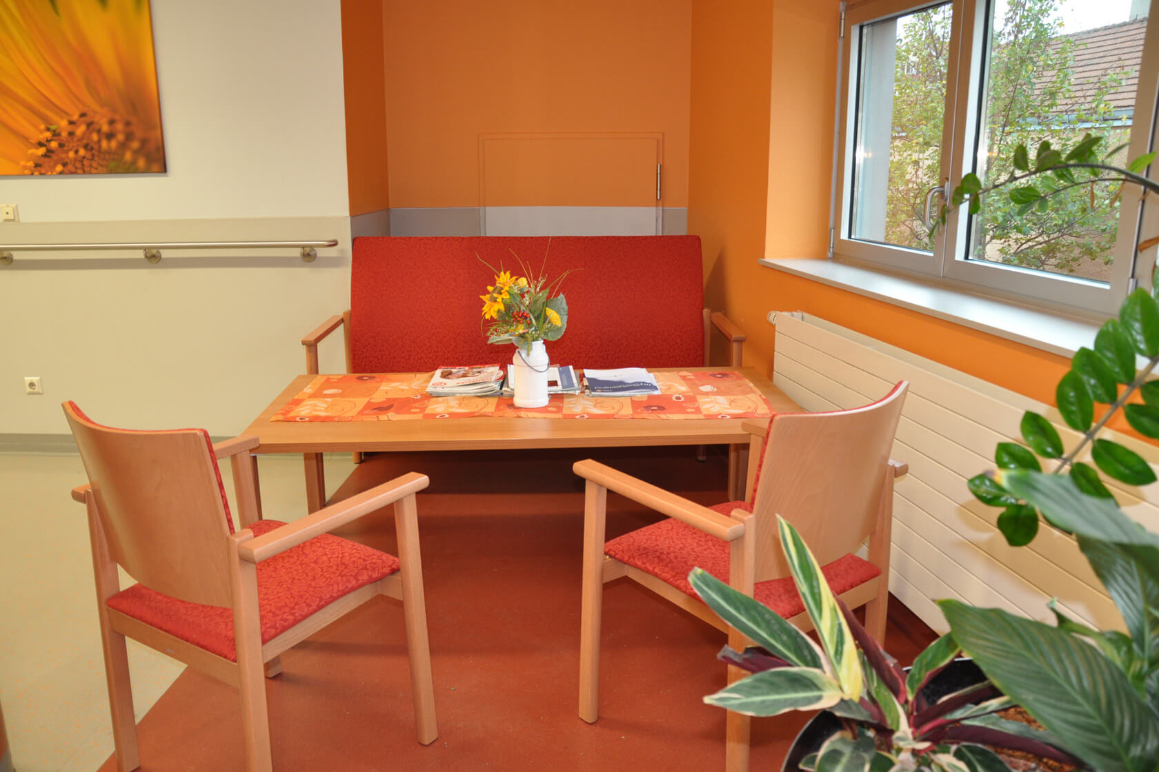 Vor einer orange gestrichenen Wand steht eine rot bezogenen Sitzbank, ein Holztisch und zwei rotbezogene Stühle.