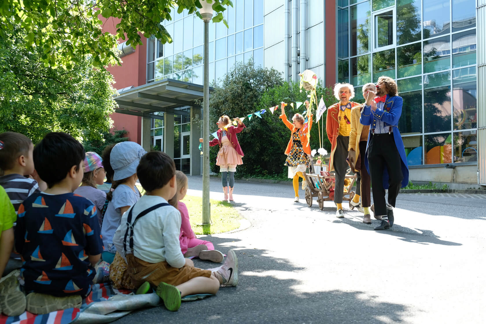 Vor einem Gebäude steht eine Gruppe als Clown verkleideter Menschen. vor ihnen sitzt eine Gruppe Kinder auf einer Wiese.