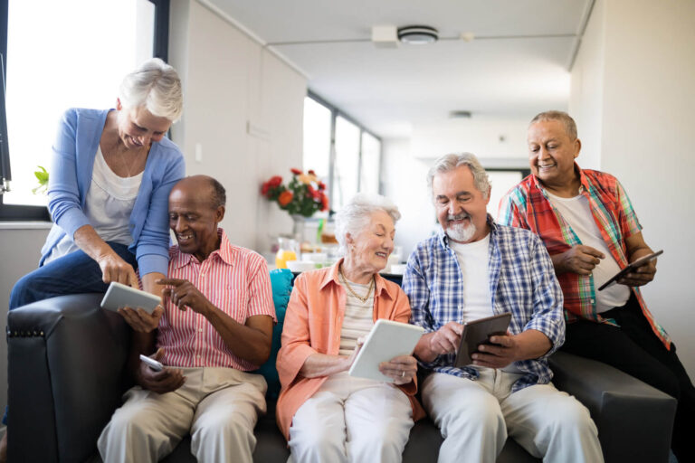 Eine Gruppe Senior:innen verschiedener Herkunft sitzt auf einen Sofa. Sie halten Tablets in den Händen, zeigen einander Dinge auf den Bildschirmen und lächeln.