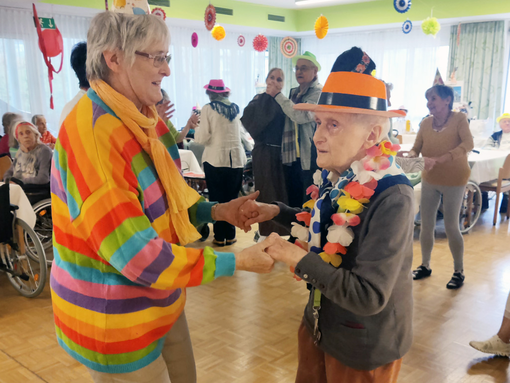 Bewohnerin im Pflegewohnheim Aigner Rollett tanzt mit Halsschmuck und Hut verkleidet mit einer Angehörigen zu Fasching