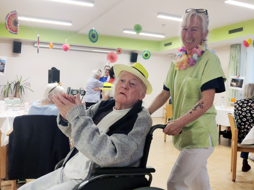 Bewohner im Pflegewohnheim Aigner Rollett klatscht zu Fasching mit Hut verkleidet zu Musik. Neben ihm steht eine Pflegefachkraft.