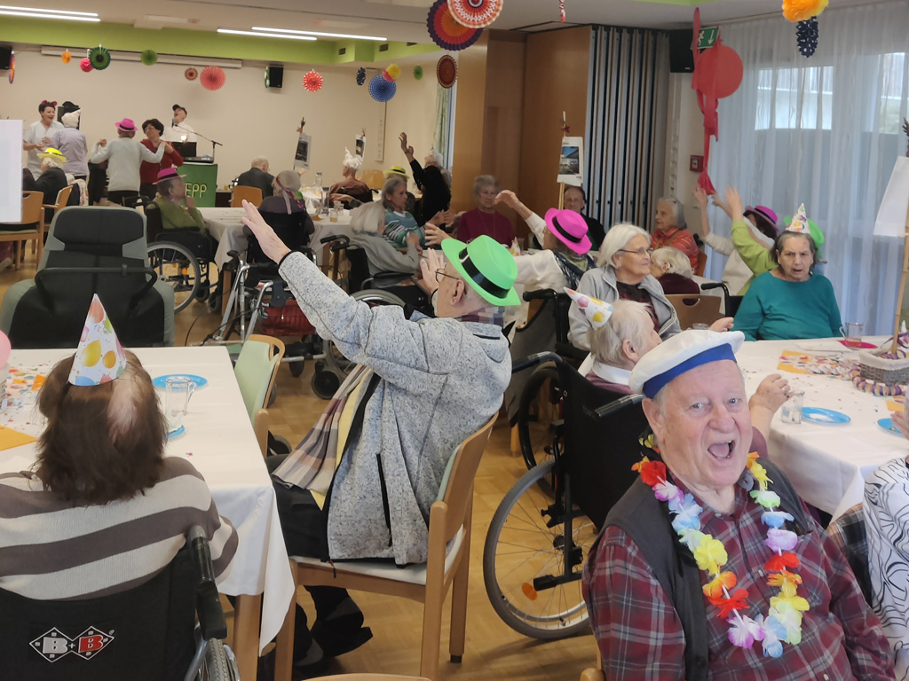 Bewohner:innen im Pflegewohnheim Aigner Rollett feiern verkleidet gemeinsam Fasching. Sie sitzen an verschiedenen Tischen und hören der Live Musik zu.