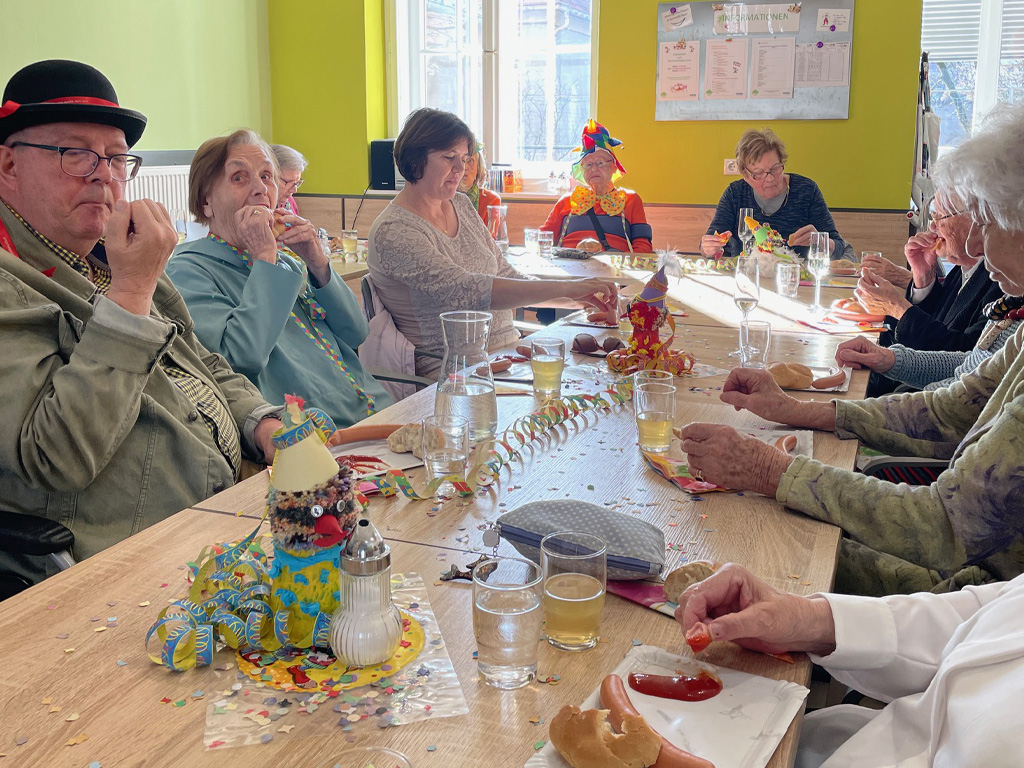 Bewohner:innen in der Wohnoase Robert Stolz sitzen am Rosenmontag gemeinsam am Tisch und essen am geschmückten Tisch Frankfurter und trinken Sekt