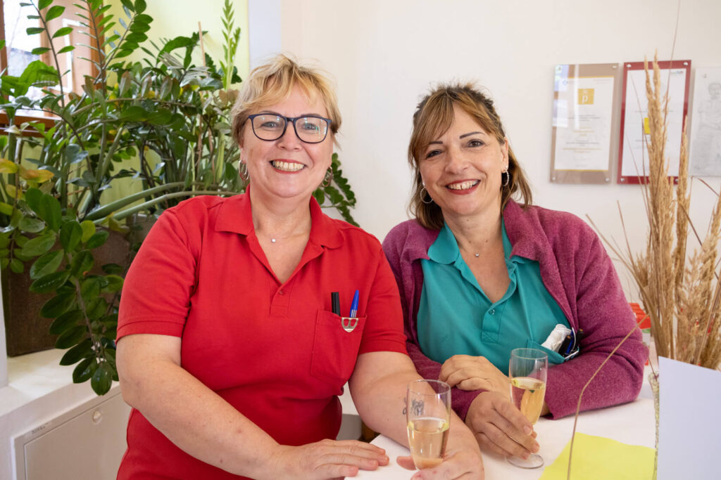 Zwei Pflegerinnen in roter und türkiser Kleidung stehen vor einem Stehtisch und lächeln.