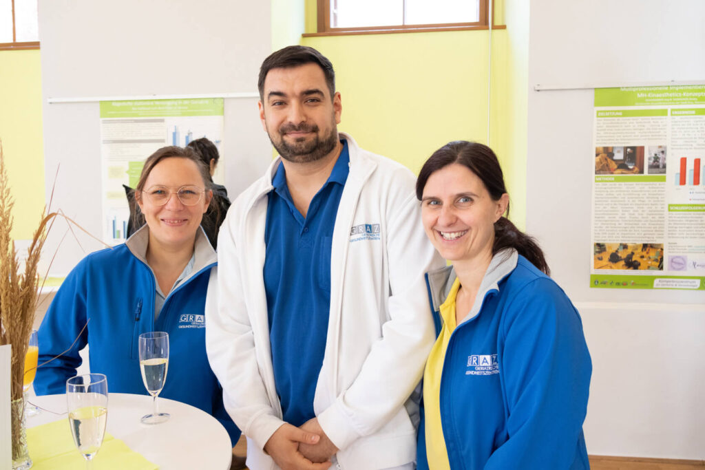 Drei Pflegekräfte in blau und weiß gekleidet stehen vor einem Stehtisch und lächeln in die Kamera.